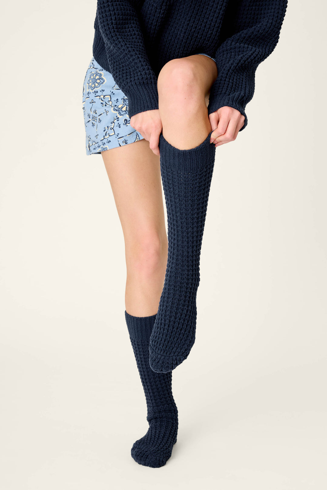 A person wearing a dark blue sweater and floral shorts pulls up a Cozy Chenille Waffle Deep Navy Mid-Calf Sock from P.J. Salvage on one leg while the other sock is already on.
