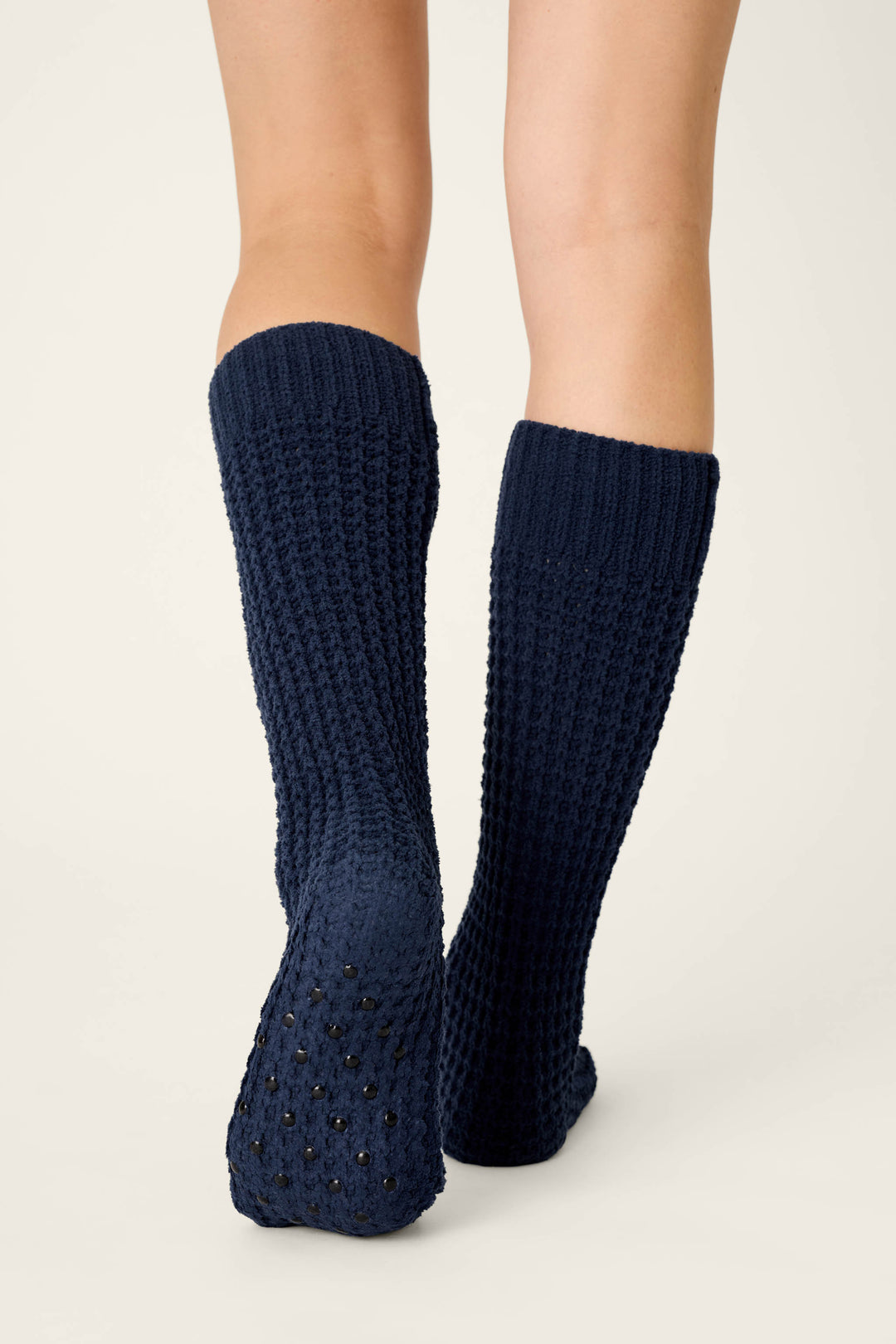 A person wearing P.J. Salvage Cozy Chenille Waffle Deep Navy Mid-Calf Socks stands on a light surface, showing the backs of their lower legs and feet. The textured socks feature rubber grip dots on the soles.