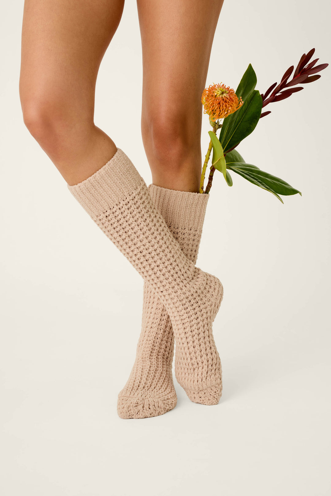 A person wearing P.J. Salvage Cozy Chenille Waffle Smoky Beige Mid-Calf Socks stands with legs crossed, holding a stem with green leaves and orange flowers behind one knee against a plain light background.