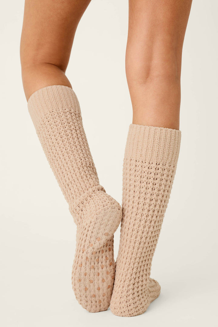 A person wearing P.J. Salvage Cozy Chenille Waffle Smoky Beige Mid-Calf Socks stands with legs crossed at the ankles, viewed from behind against a plain light background.