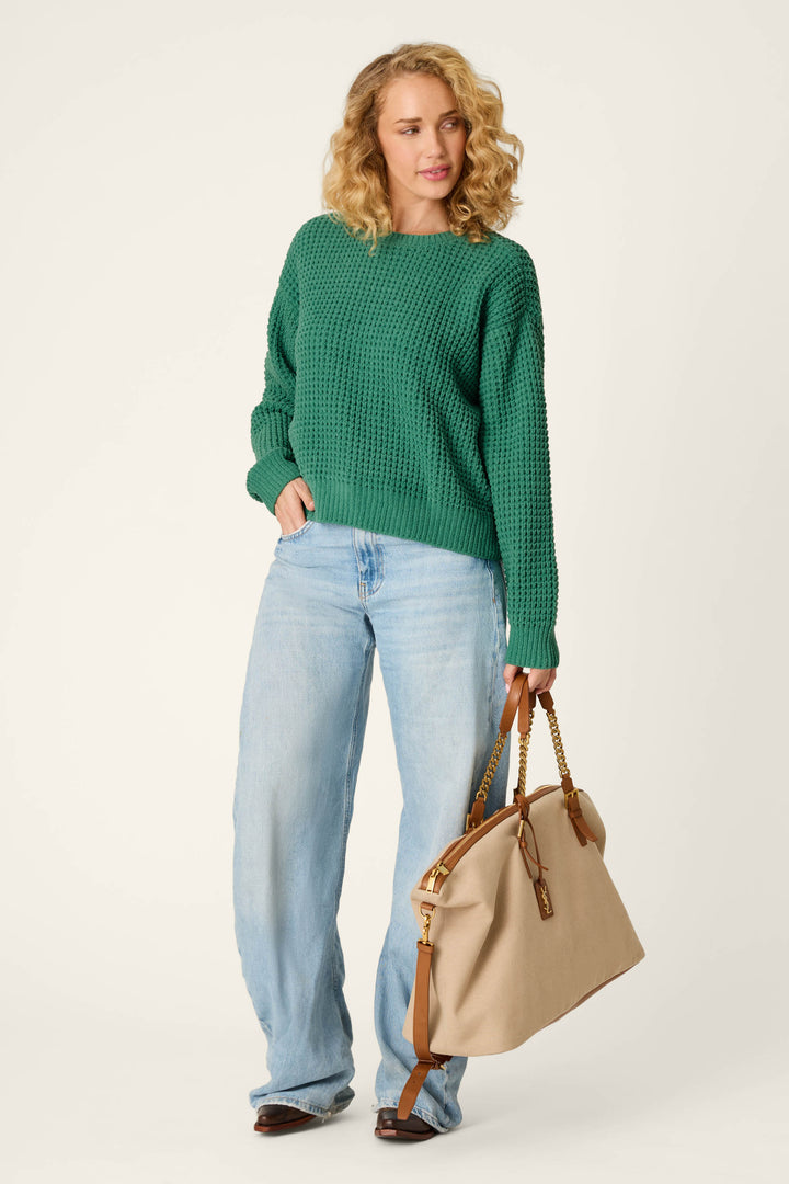 A woman with curly blonde hair wears the P.J. Salvage Cozy Chenille Waffle Green Sweater and light blue jeans, standing with a large beige handbag with brown handles, looking to her left against a plain background.