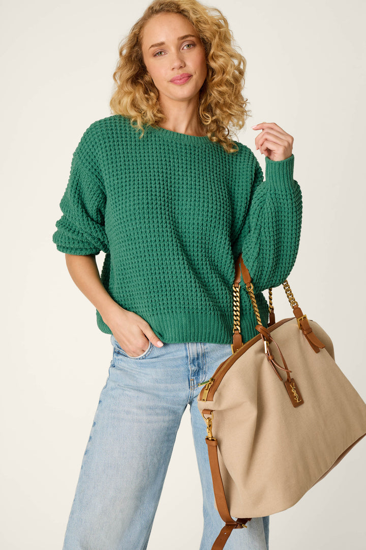 A woman with curly blonde hair wears the Cozy Chenille Waffle Green Sweater by P.J. Salvage and light blue jeans, holding a large beige and brown handbag with chain straps, standing against a plain light background.