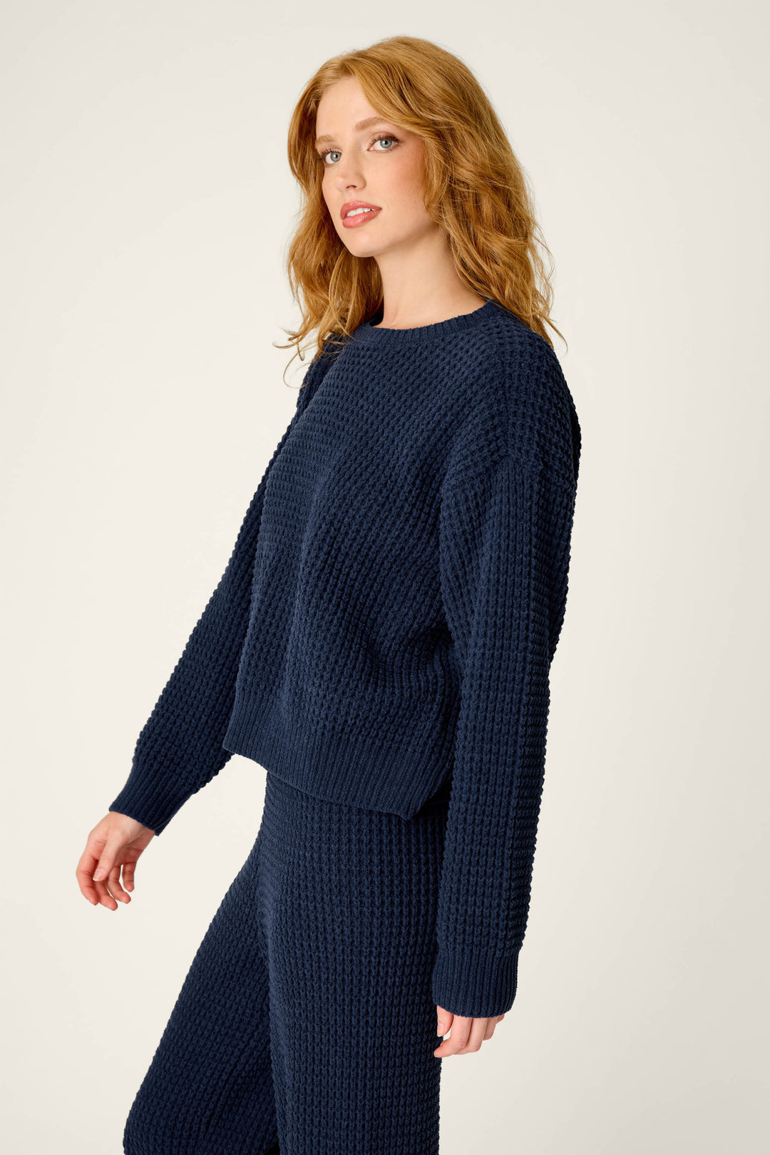 A woman with wavy red hair wears the P.J. Salvage Cozy Chenille Waffle Deep Navy Sweater, paired with matching pants, standing against a plain light background and looking slightly to the side.