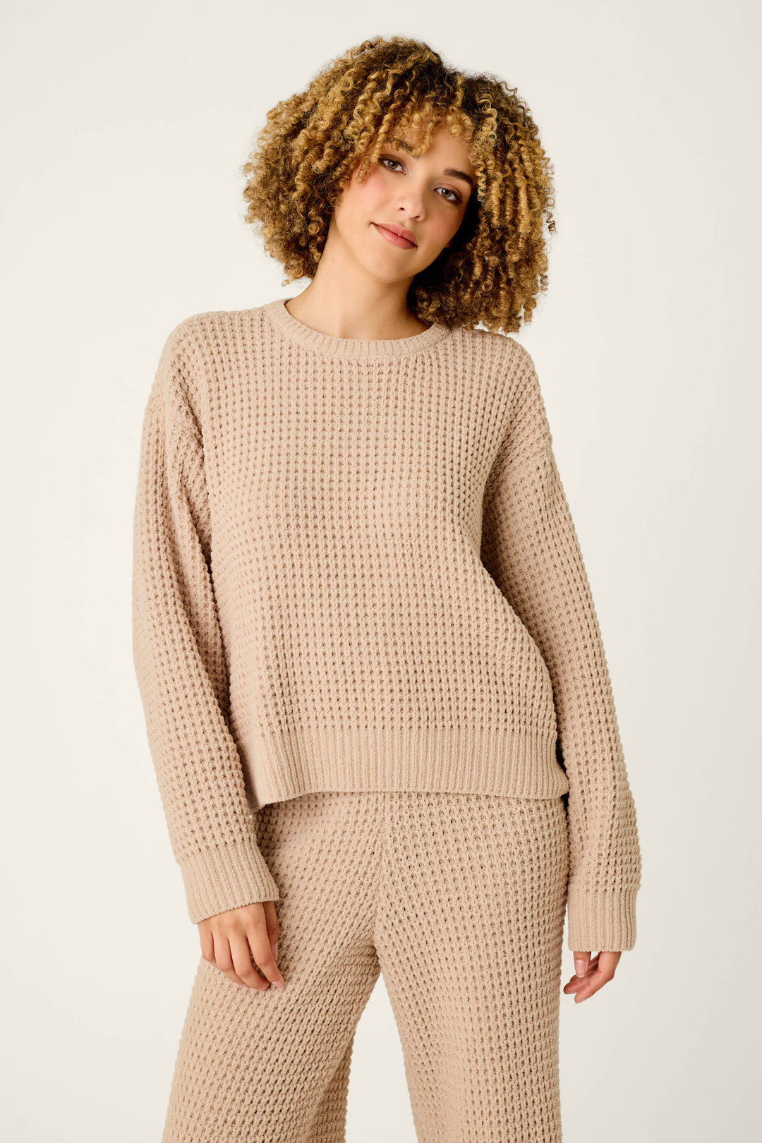 Someone with curly hair is wearing the Cozy Chenille Waffle Smoky Beige Sweater by P.J. Salvage along with matching pants, standing against a plain light background and smiling softly.