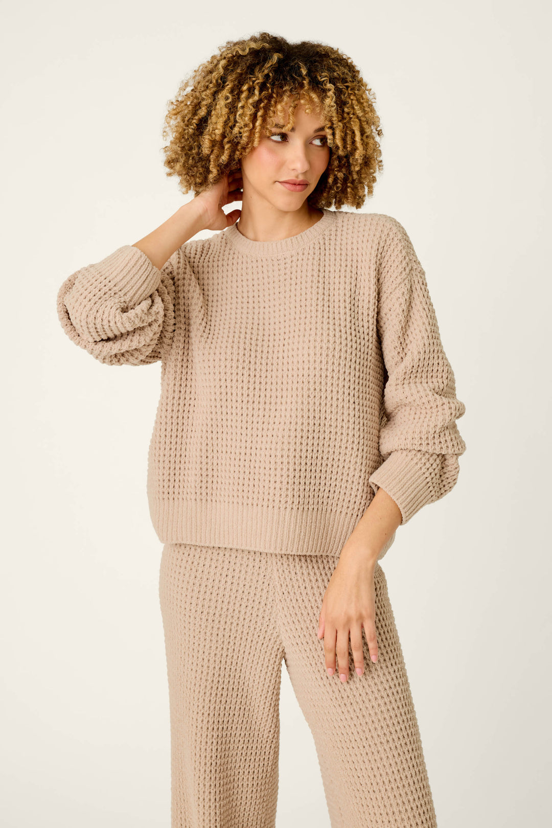 A woman with curly hair wears the P.J. Salvage Cozy Chenille Waffle Smoky Beige Sweater, standing against a plain, light background, touching her neck and gazing to the side.
