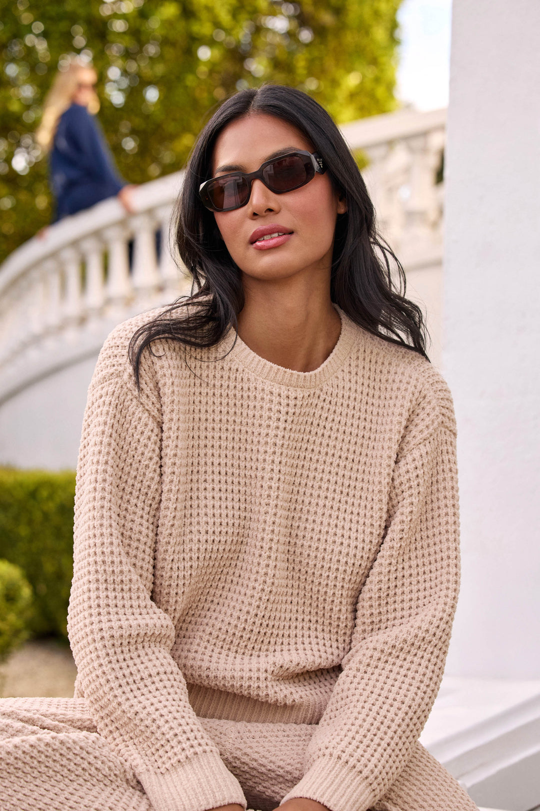 A woman with long dark hair, wearing sunglasses and the P.J. Salvage Cozy Chenille Waffle Smoky Beige Sweater, sits outdoors near a white wall and greenery. Another person is blurred in the background on a curved stairway.