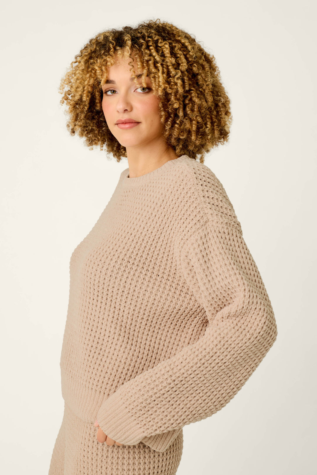 A person with curly hair wears the P.J. Salvage Cozy Chenille Waffle Smoky Beige Sweater, paired with matching pants, standing against an off-white background and gazing at the camera with a soft expression.