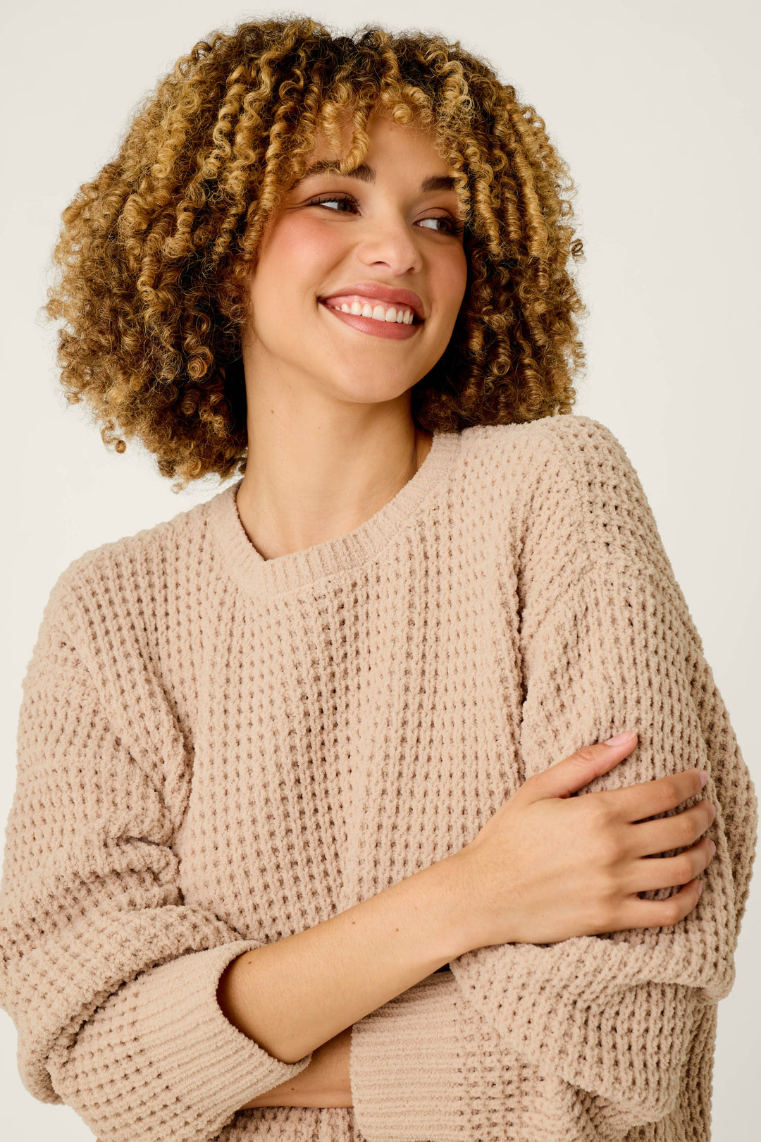 A smiling woman with curly hair wears the P.J. Salvage Cozy Chenille Waffle Smoky Beige Sweater, crossing her arms and glancing to the side against a plain light background.