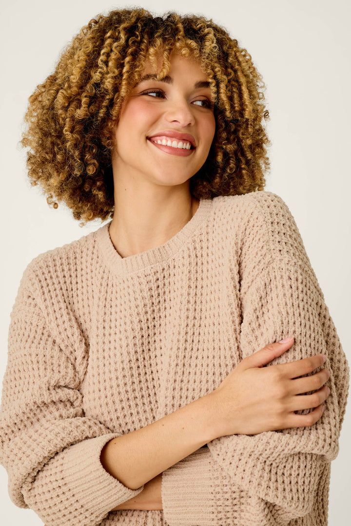 A smiling woman with curly hair wears the P.J. Salvage Cozy Chenille Waffle Smoky Beige Sweater, crossing her arms and glancing to the side against a plain light background.