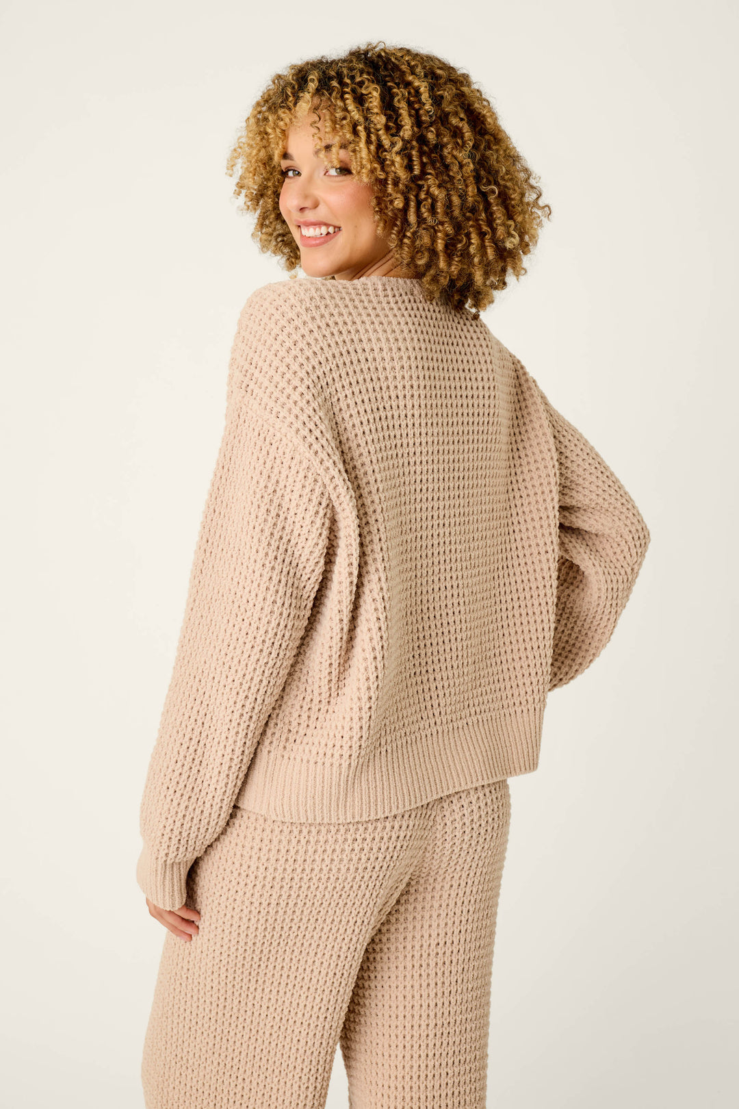 A woman with curly hair smiles over her shoulder, wearing the P.J. Salvage Cozy Chenille Waffle Smoky Beige Sweater, standing against a plain, light background.