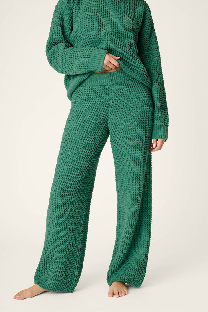 A person models the P.J. Salvage Cozy Chenille Waffle Green Lounge Pants, paired with a loose, textured green knit sweater. Barefoot against a plain light background, they gently lift the hem of the sweater with their left hand, showcasing a comfortable and cozy style.