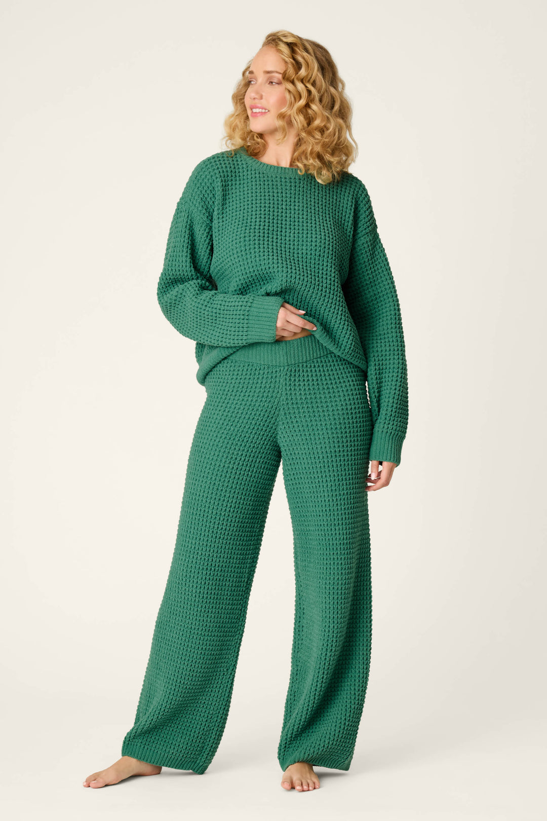 A woman with wavy blonde hair wears the Cozy Chenille Waffle Green Lounge Pants by P.J. Salvage, along with a matching loose, textured green sweater. She stands barefoot on a light background, looking off to the side and smiling.
