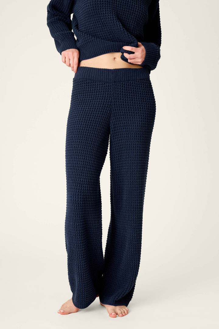 A person is wearing P.J. Salvage’s Cozy Chenille Waffle Deep Navy Lounge Pants with a matching long-sleeve top, barefoot and gently lifting the sweater to reveal their midriff.