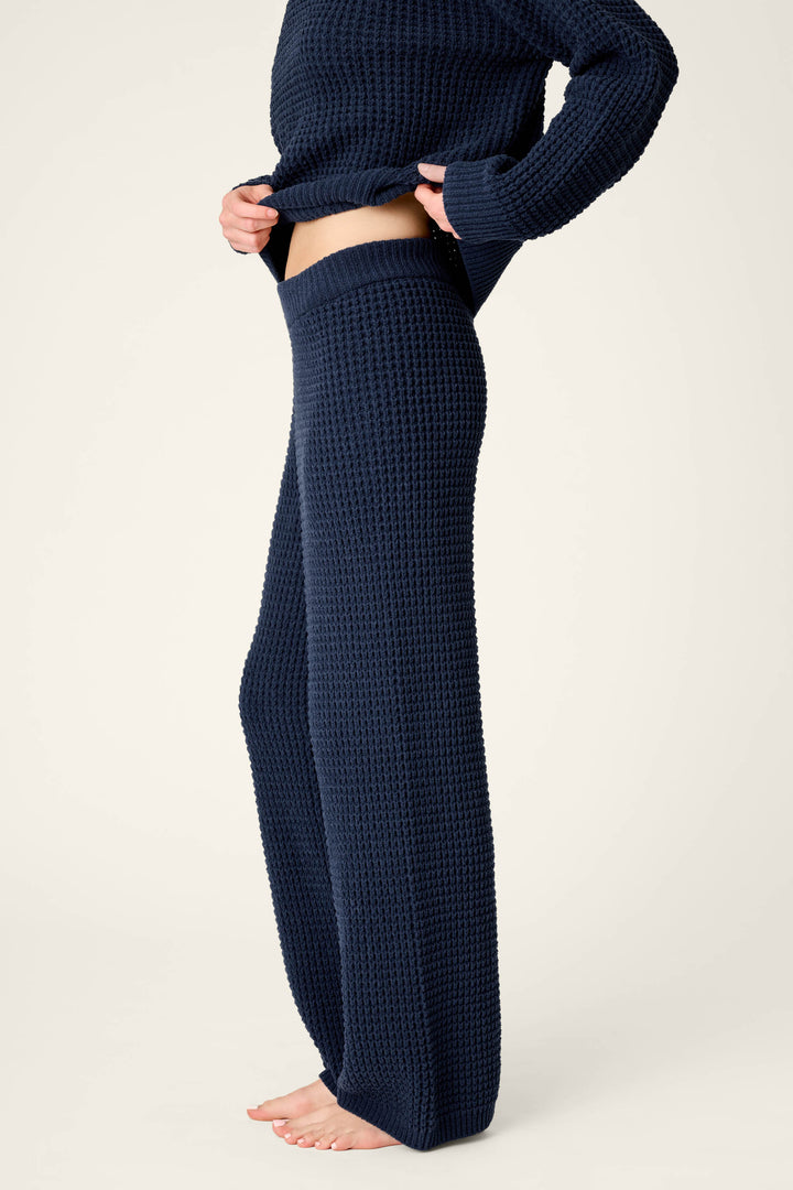 A person is barefoot on a light background, slightly lifting their sweater to reveal the waistband of their Cozy Chenille Waffle Deep Navy Lounge Pants by P.J. Salvage, paired with a matching dark blue top.