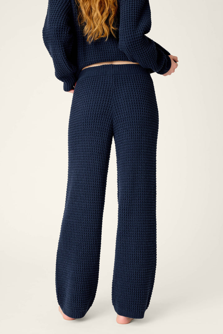 A person with long, wavy, reddish-blonde hair is shown from the back, standing barefoot against a plain light-colored background. They are wearing the P.J. Salvage Cozy Chenille Waffle Deep Navy Lounge Pants and a matching long-sleeve top.