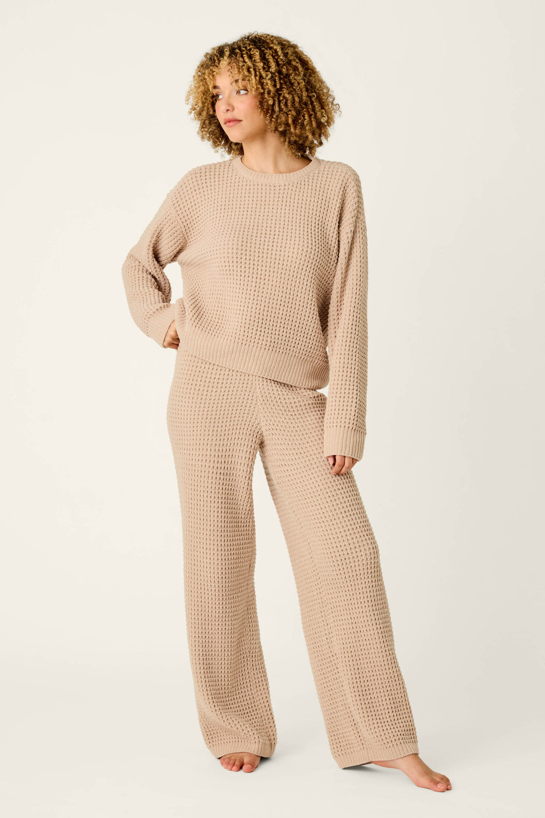 A woman with curly hair stands barefoot against a plain light background, wearing P.J. Salvage’s Cozy Chenille Waffle Smoky Beige Lounge Pants and a matching loose sweater.