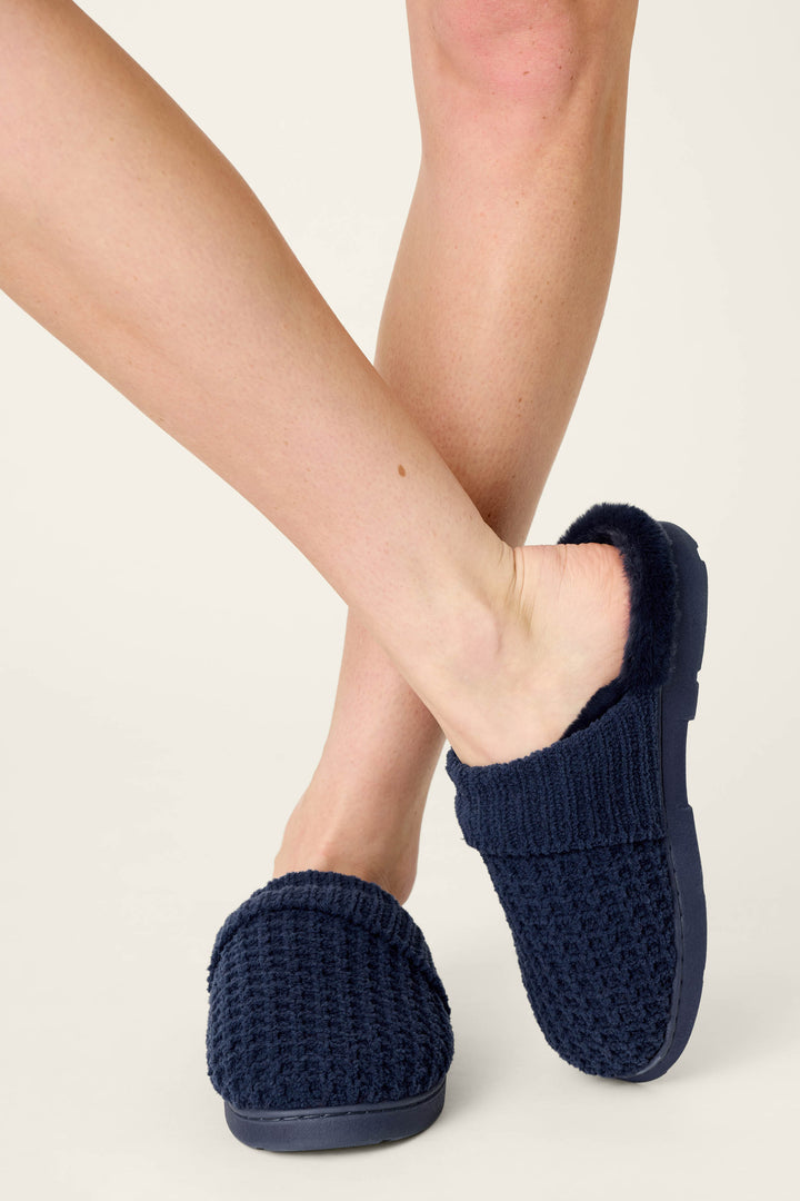 A person stands with legs crossed, wearing P.J. Salvage Cozy Chenille Waffle Deep Navy Slippers with a fuzzy lining on a light background.