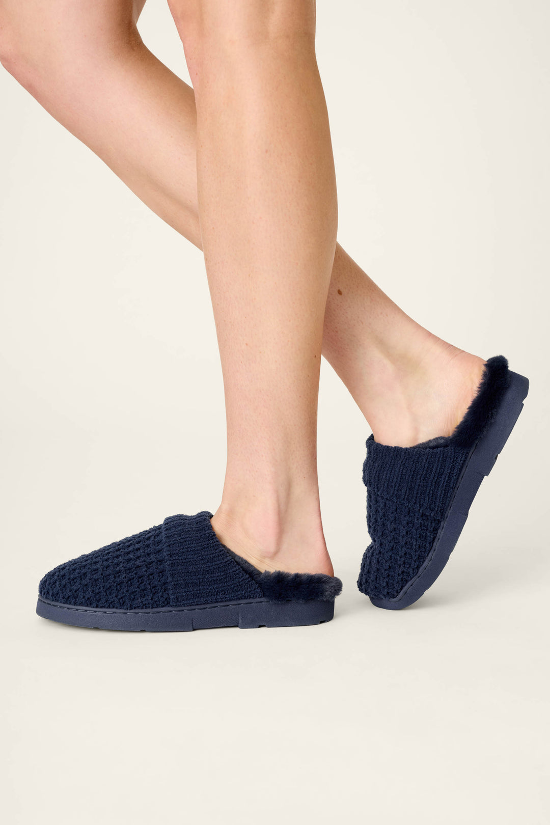 A person wears P.J. Salvage Cozy Chenille Waffle Deep Navy Slippers, featuring a knit-textured design and fuzzy lining, while standing on a light background with one foot slightly lifted behind the other.
