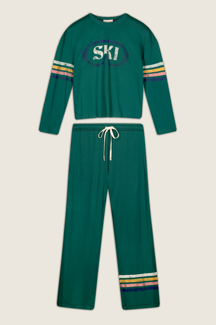 The P.J. Salvage Ski Goggles Graphic Long Sleeve Pajama Set is a green two-piece loungewear set featuring a long-sleeve top with “SKI” lettering and striped accents on the sleeves, while the matching drawstring pants have coordinating stripes near the cuffs. Both pieces are finished with yellow, white, and purple accents for a stylish touch.