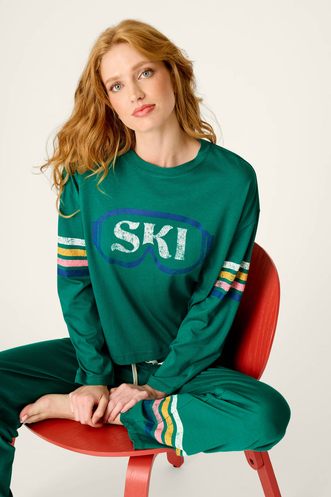 A woman with long red hair sits on a red chair, barefoot and relaxed, wearing the P.J. Salvage Ski Goggles Graphic Long Sleeve Pajama Set, which features a green top with SKI text on the front and colorful striped pants.