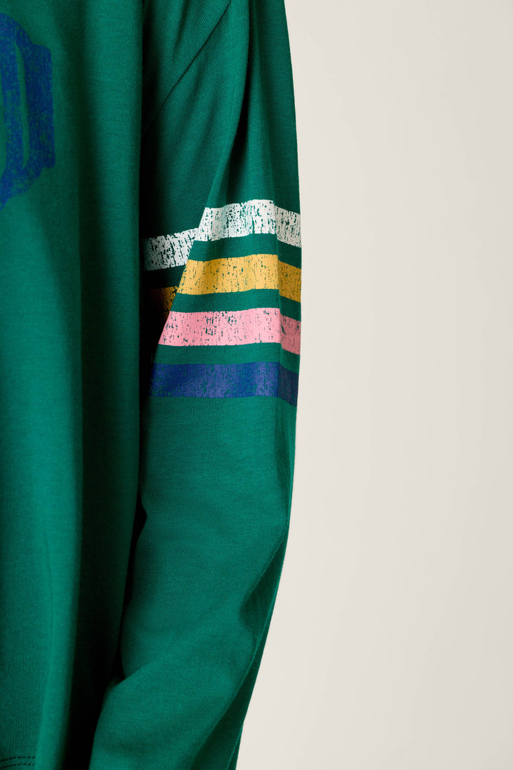 A long-sleeve pajama top from P.J. Salvage’s Ski Goggles Graphic Long Sleeve Pajama Set, featuring green fabric with horizontal stripes in white, yellow, green, pink, and blue on the upper arm. The background is plain and light-colored.