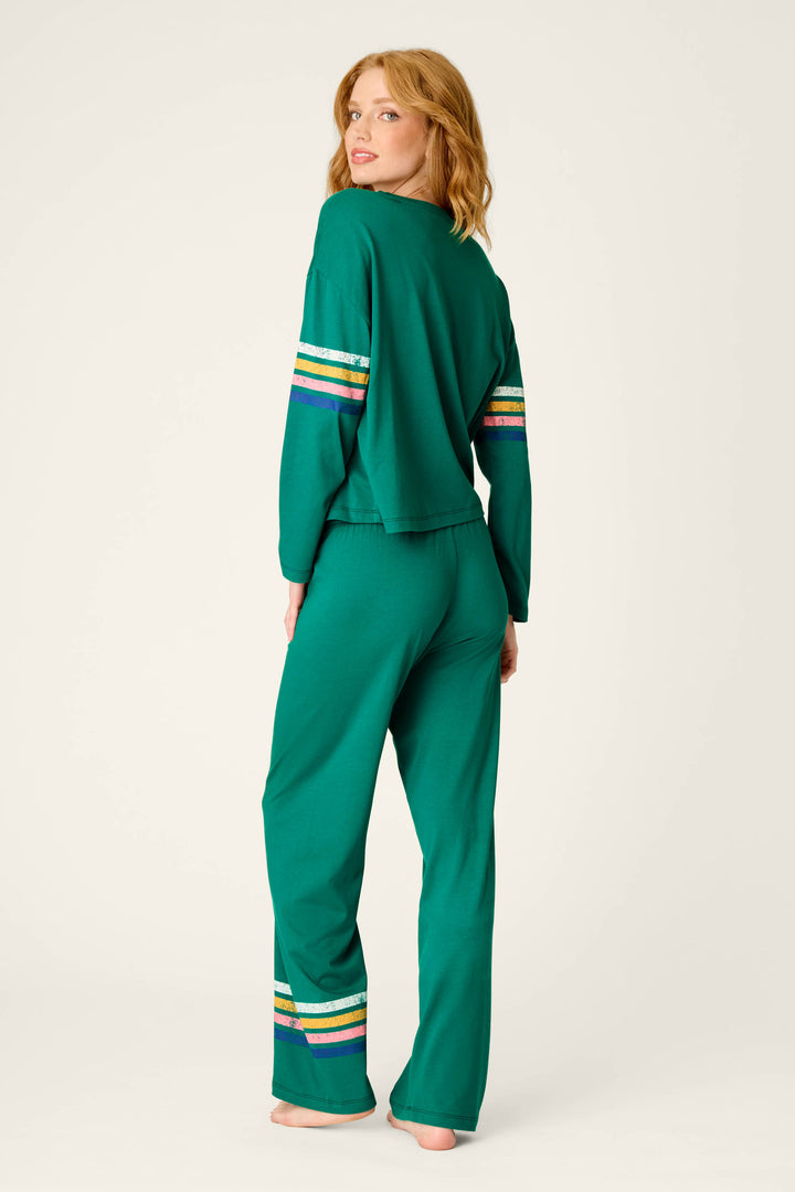 A woman in the P.J. Salvage Ski Goggles Graphic Long Sleeve Pajama Set, featuring striped details on the sleeves and pants, stands barefoot and looks over her shoulder against a plain background.