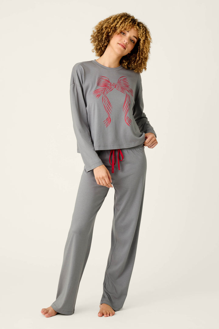 A woman with curly hair models the P.J. Salvage Bow Graphic Long Sleeve Pajama Set, featuring gray pajamas adorned with a red bow design on the top and red drawstrings on the pants. She stands barefoot against a light, plain background, one hand in her pocket and posing in a relaxed stance.