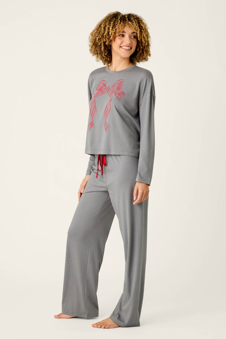 A woman models the P.J. Salvage Bow Graphic Long Sleeve Pajama Set, featuring loose gray pajamas with a red bow design on the top and red drawstring pants. She stands barefoot on a neutral background, smiling and looking to the side.