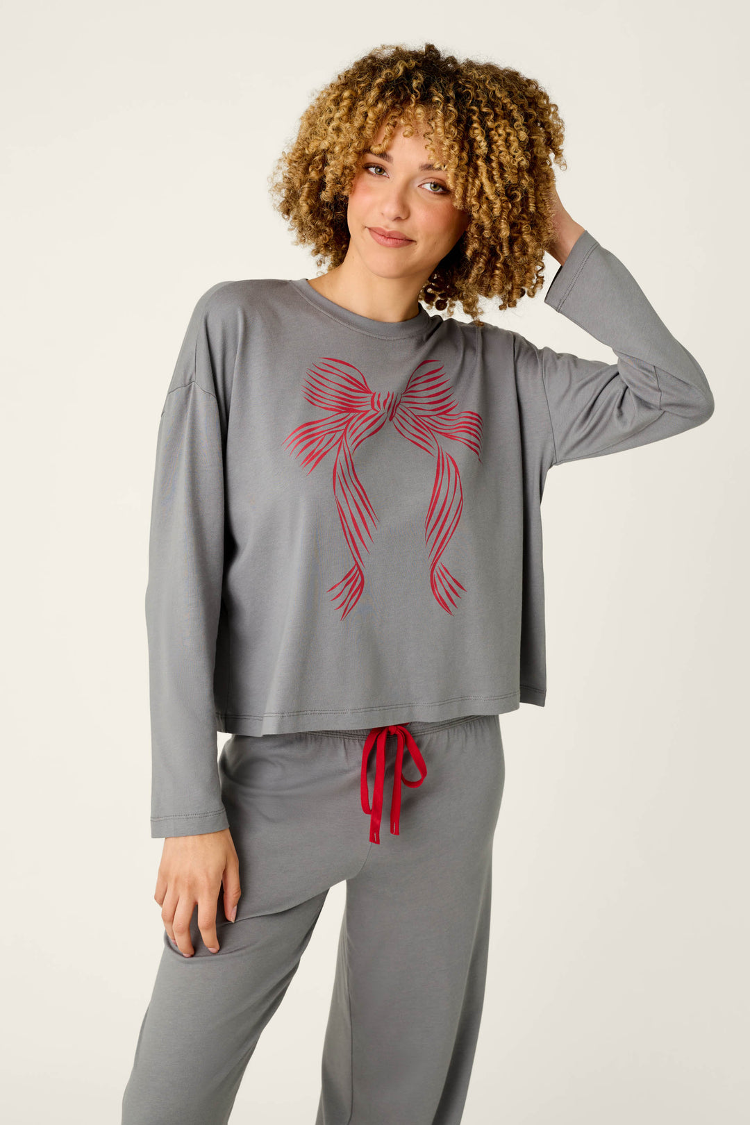 A woman with curly hair stands casually, touching her hair, dressed in the P.J. Salvage Bow Graphic Long Sleeve Pajama Set—a loose gray ensemble featuring a bold red bow graphic on the top and matching red drawstring on the pants.
