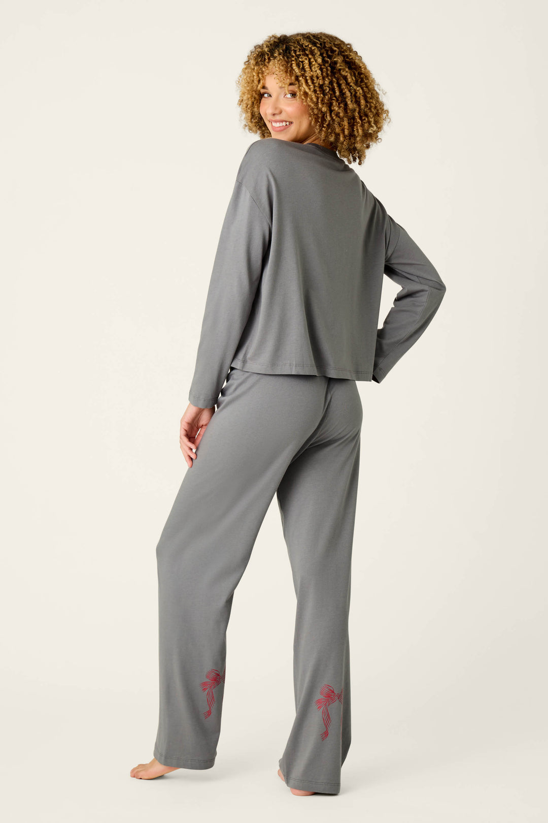 A woman with curly hair smiles and poses barefoot against a plain background, wearing the P.J. Salvage Bow Graphic Long Sleeve Pajama Set, a matching gray outfit featuring subtle red bow designs near the ankles.