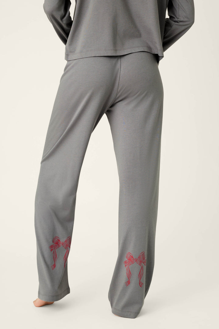 A person is shown from behind, barefoot and wearing the P.J. Salvage Bow Graphic Long Sleeve Pajama Set, featuring gray pajama pants with red bow designs on the lower legs.