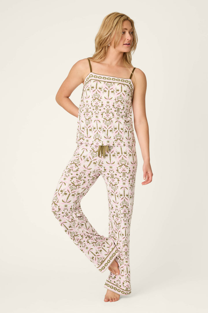 A woman wears the P.J. Salvage Déjà Vu Dreams Tiki Time Long PJ Set, a matching white pajama set with pink and green florals, sleeveless top, and long pants. She stands barefoot against a plain, light background.