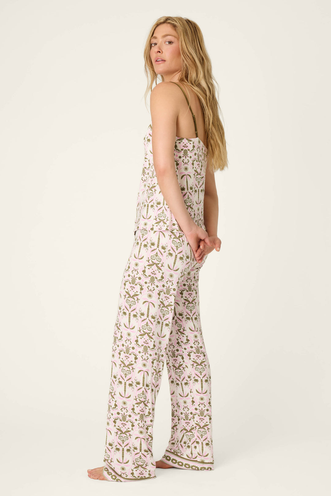 A person with long blonde hair stands barefoot, looking over their shoulder, wearing the Déjà Vu Dreams Tiki Time Long PJ Set by P.J. Salvage—a matching sleeveless top and wide-leg pants with a pink and green floral print on white.