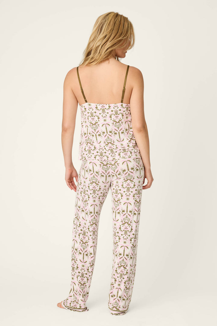 A woman with shoulder-length blonde hair stands facing away, wearing the P.J. Salvage Déjà Vu Dreams Tiki Time Long PJ Set—a white, floral pajama set featuring pink and green patterns, a spaghetti strap top, and loose-fitting pants.