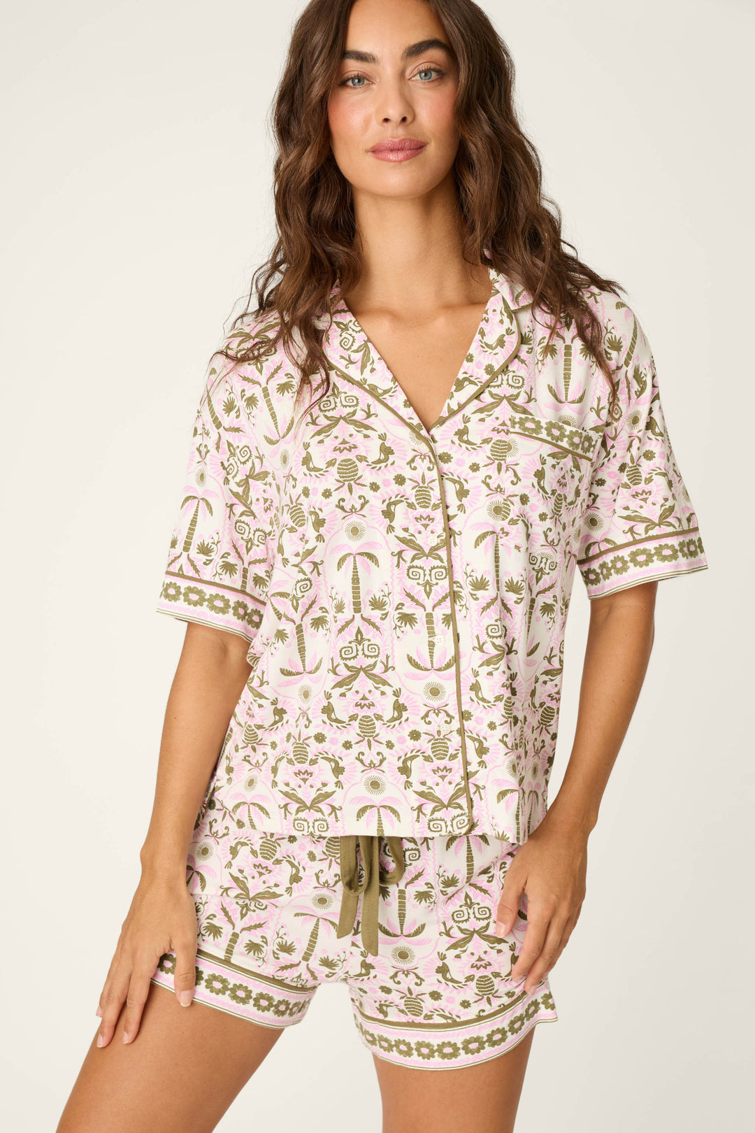 A woman with wavy brown hair smiles at the camera, wearing the P.J. Salvage Déjà Vu Dreams Tiki Time Short Sleeve Top—a matching button-up pajama top and drawstring shorts with a pink and green floral pattern.