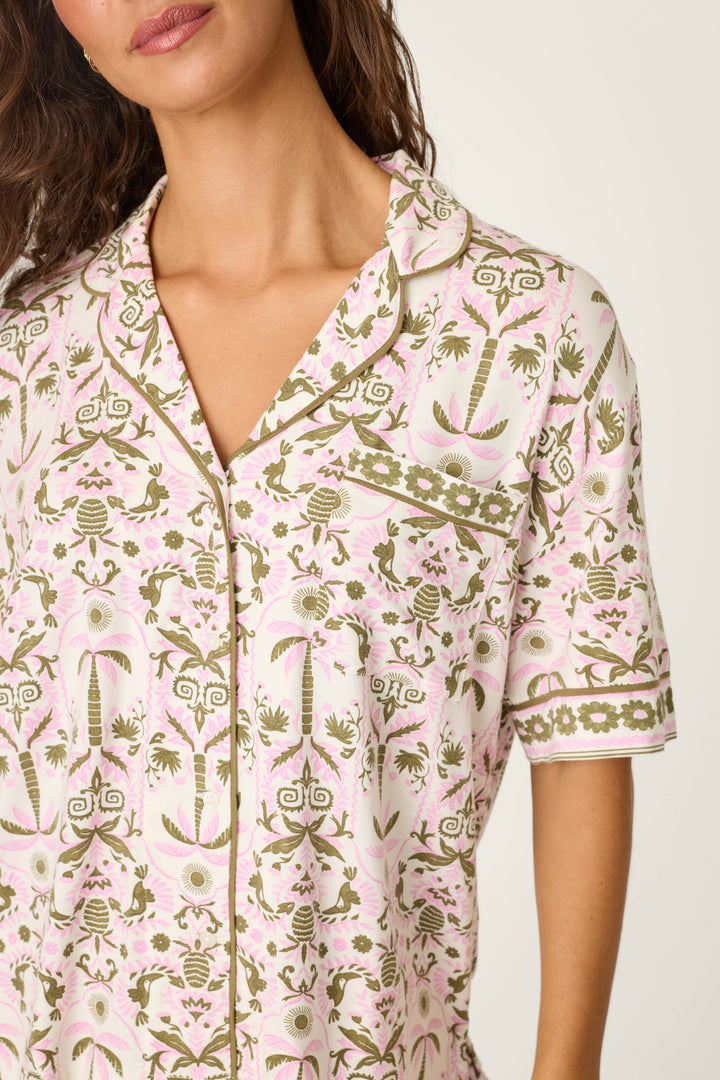 A woman wears the P.J. Salvage Déjà Vu Dreams Tiki Time Short Sleeve Top, featuring light pink and white fabric with green and pink floral and geometric patterns, a collar, buttons, short sleeves, and a chest pocket. Image cropped at chin and mid-torso.