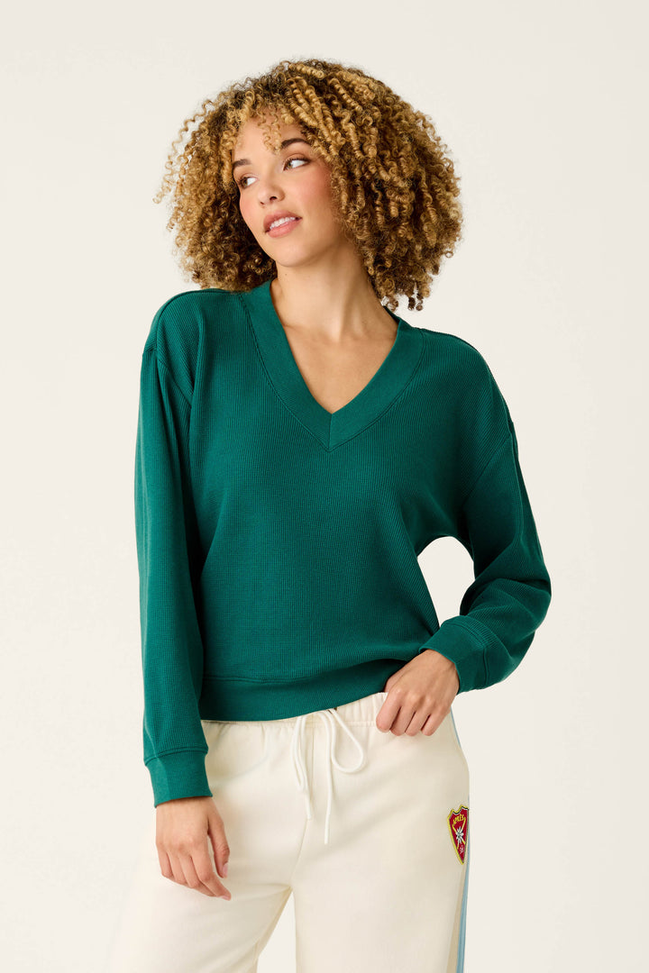 A woman with curly hair wears the Weekday Waffles Deep Dive Dark Green Long Sleeve Top by P.J. Salvage and white pants with a small red patch, standing against a plain light background and looking to the side.
