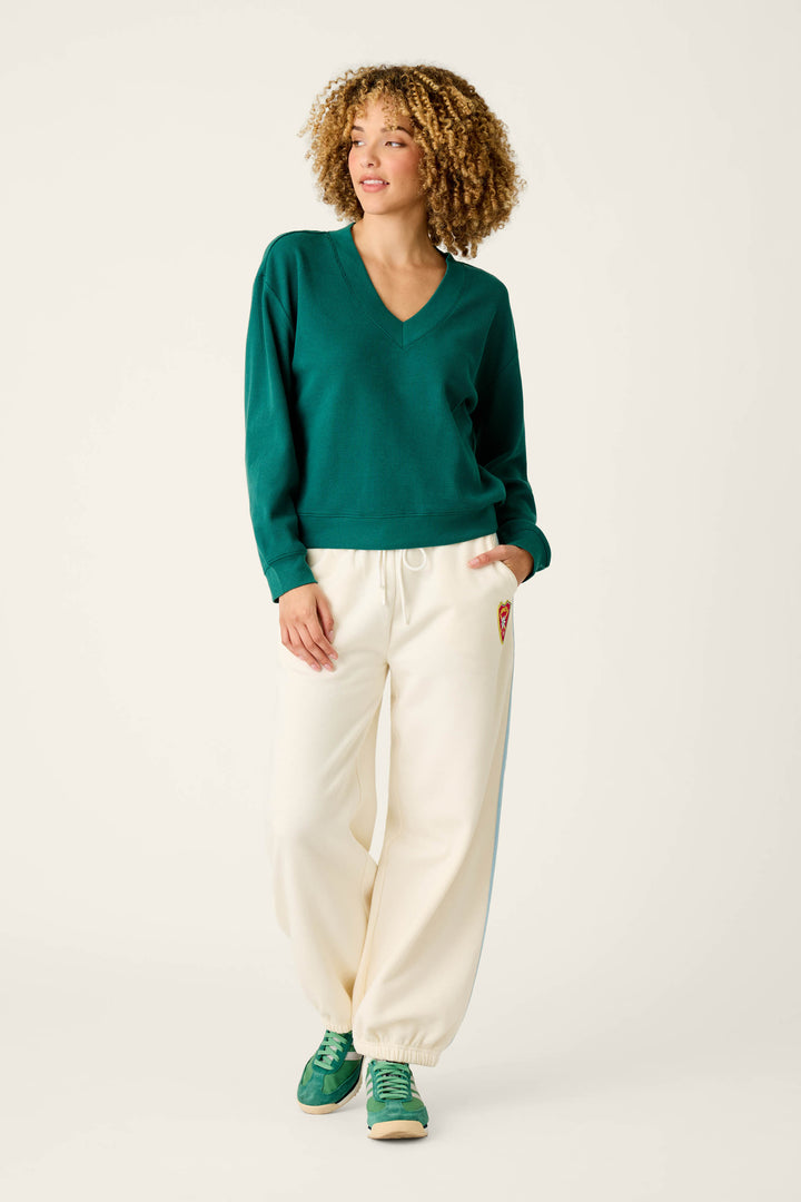 A person with curly hair stands with one hand in their pocket against a plain background, wearing the Weekday Waffles Deep Dive Dark Green Long Sleeve Top by P.J. Salvage, paired with white drawstring sweatpants featuring a small patch and green sneakers.