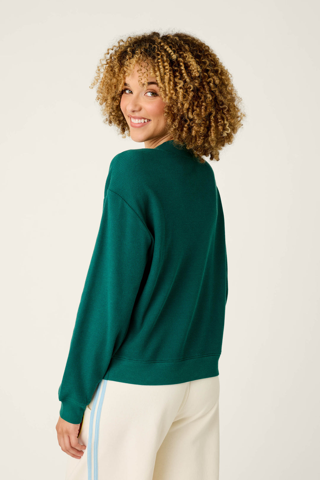 Wearing the P.J. Salvage Weekday Waffles Deep Dive Dark Green Long Sleeve Top, a person with curly hair and cream pants with blue stripes looks back over their shoulder and smiles against a plain light background.