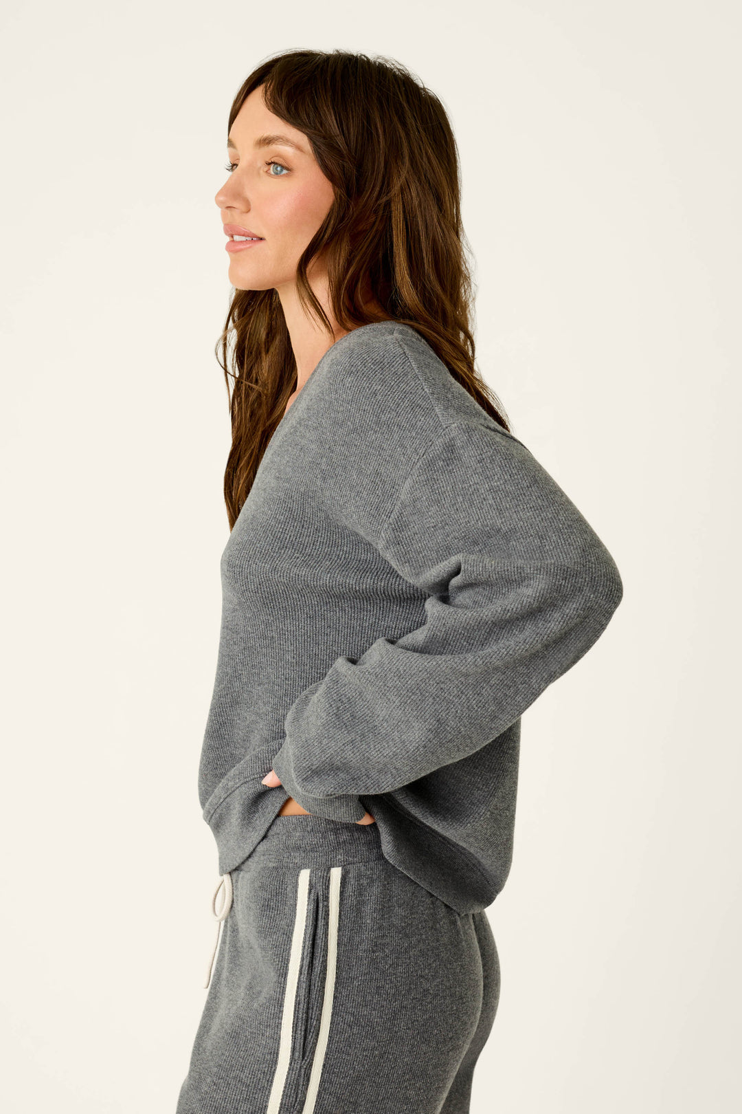 A woman with long brown hair is standing in profile against a plain light background, wearing the P.J. Salvage Weekday Waffles Deep Dive Charcoal Long Sleeve Top—a loose-fitting gray sweater—paired with matching gray sweatpants featuring white stripes.