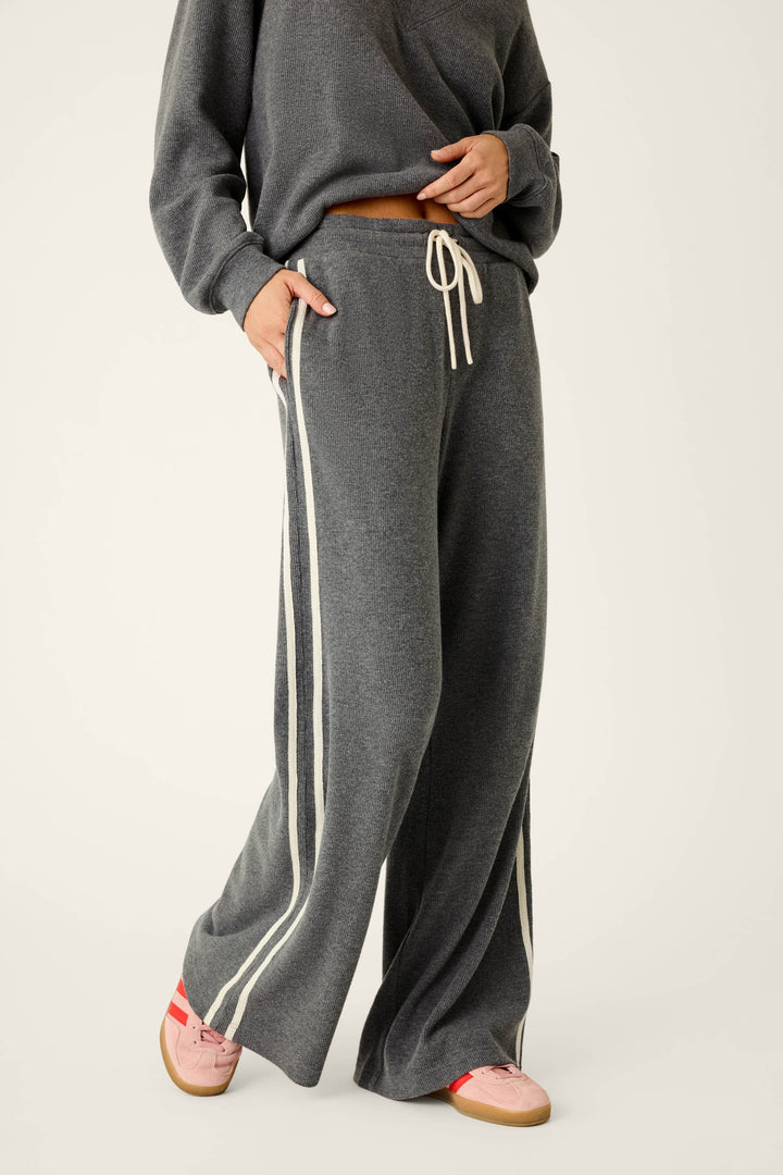 Wearing the P.J. Salvage Weekday Waffles Honey Charcoal Lounge Pants with white side stripes and a matching gray sweatshirt, this person stands with one hand in their pocket. Pink and tan sneakers complete the look against a plain, light-colored background.