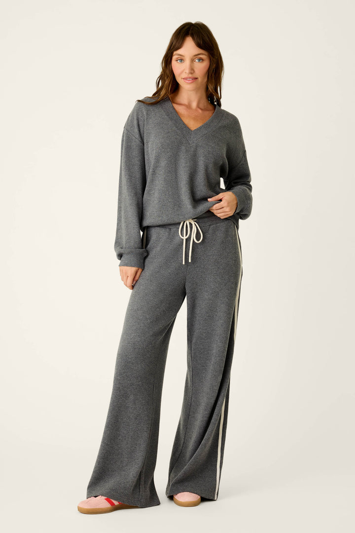 A woman stands and smiles against a plain background, wearing the P.J. Salvage Weekday Waffles Honey Charcoal Lounge Pants—loose-fitting, wide-leg pants with a white drawstring and side stripe—paired with a matching grey V-neck sweater.