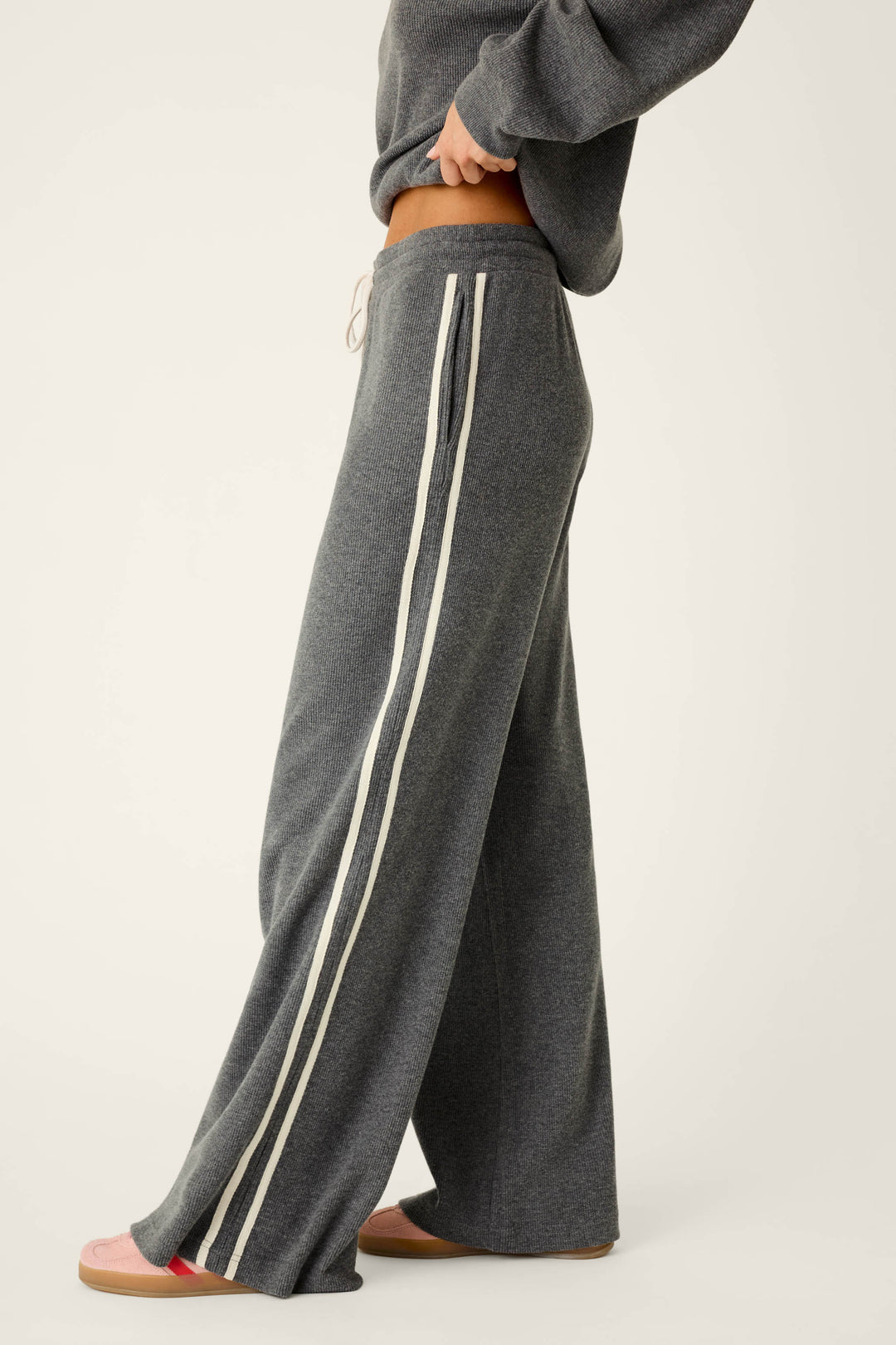 A person is wearing the P.J. Salvage Weekday Waffles Honey Charcoal Lounge Pants—loose-fitting gray pants with white side stripes—paired with a matching gray sweatshirt and pink slippers, standing sideways against a plain light background.