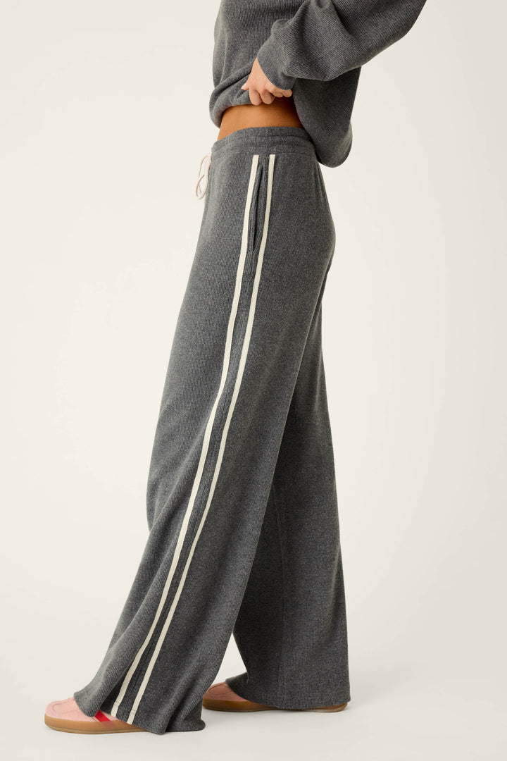 A person is wearing the P.J. Salvage Weekday Waffles Honey Charcoal Lounge Pants—loose-fitting gray pants with white side stripes—paired with a matching gray sweatshirt and pink slippers, standing sideways against a plain light background.