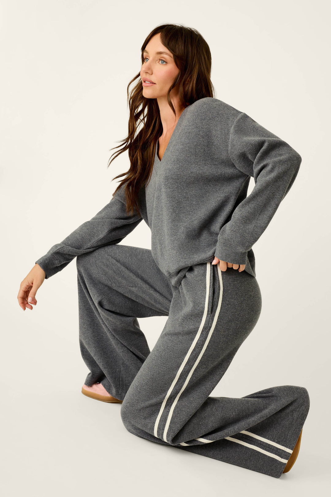 A woman with long brown hair kneels on one knee, wearing the P.J. Salvage Weekday Waffles Honey Charcoal Lounge Pants, a pair of loose, wide-leg pants with white side stripes, along with a coordinating gray sweater, against a plain light background.