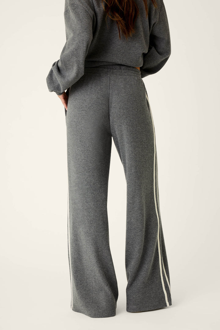 A person stands facing away against a plain background, showcasing the back and lower torso while wearing P.J. Salvage’s Weekday Waffles Honey Charcoal Lounge Pants, which feature gray wide legs and white side stripes.
