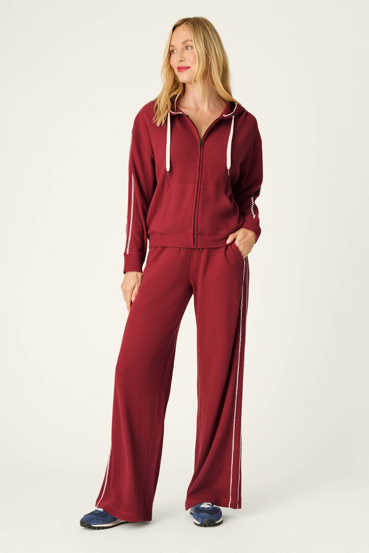 A woman stands against a plain light background, smiling to the side with one hand in her pocket. She wears the P.J. Salvage Weekday Waffles Rue Lounge Pants in maroon, paired with a matching zip-up hoodie and white sneakers.