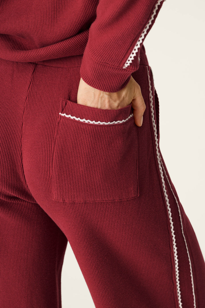 A person wearing P.J. Salvage Weekday Waffles Rue Lounge Pants in ribbed maroon with white trim has their hand in the back pocket. The matching top also features white trim along the sleeve.