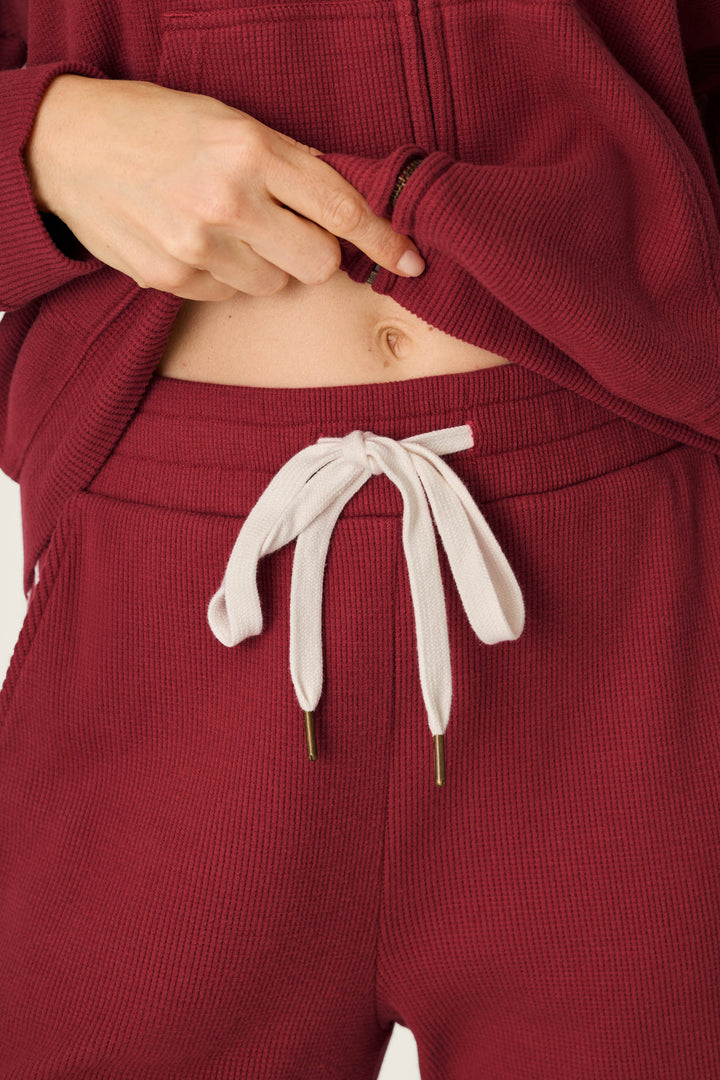 A person wearing the P.J. Salvage Weekday Waffles Rue Lounge Pants in burgundy pulls up their hoodie to reveal their belly button, displaying a white drawstring tied in a bow on the pants.