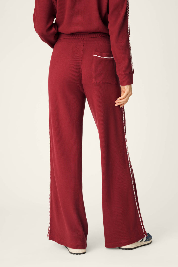 A person is seen from the back wearing P.J. Salvage’s Weekday Waffles Rue Lounge Pants in maroon, featuring white stitching and a side stripe. A matching maroon top and white sneakers are partially visible.